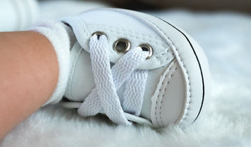 A detailed view of a white baby sneaker with laces, resting on a fluffy surface.