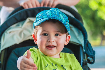 Happy child reaching out while sitting in a stroller, wearing a blue cap and green shirt, outdoors.